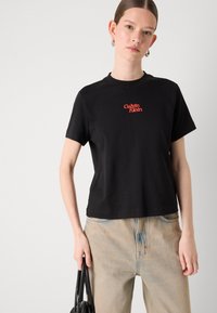Svart bomull T-skjorte med korte ermer og rund hals. Har rødt "Calvin Klein"-logo på brystet. Brukt med lyse denimbukser.