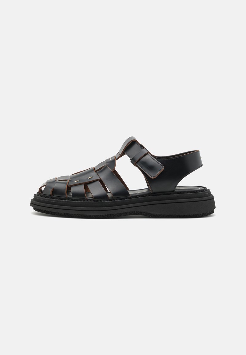 Sandales en cuir noir avec un design tissé, dotées de brides ajustables, d'accents métalliques et d'une épaisse semelle noire pour la stabilité.