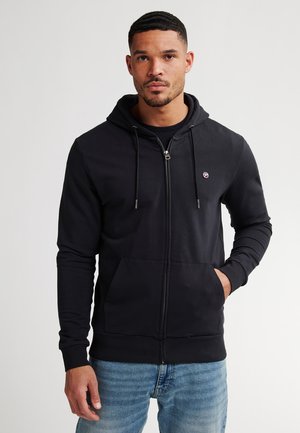 Zwarte rits hoodie met trekkoordkap, voorzakken en geribbelde manchetten. Zacht materiaal; minimale branding op de borst. Verkocht met blauwe spijkerbroeken.