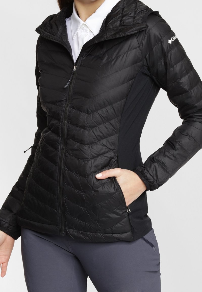 Femme portant une veste matelassée noire zippée avec des panneaux latéraux, main dans la poche, chemise blanche à col en dessous et pantalon gris.
