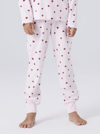 Enfant portant un pyjama rose pâle orné de motifs en forme de cœur rouge, debout pieds nus devant un fond clair.