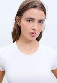 Witte getailleerde t-shirt met korte mouwen, ronde halslijn en een gladde textuur, gepresenteerd tegen een lichte achtergrond.