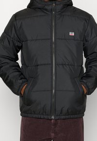 Svart vadderad jacka med huva, dragkedja framtill, justerbara manschetter och elastisk midja. Har en liten märkeslapp på bröstet.