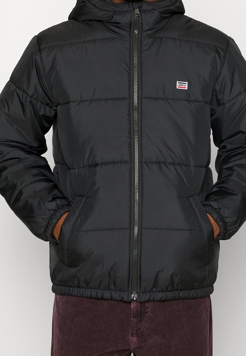 Svart vadderad jacka med huva, dragkedja framtill, justerbara manschetter och elastisk midja. Har en liten märkeslapp på bröstet.