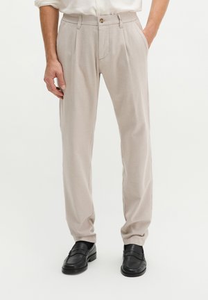 Man draagt lichtbeige rechte broek met voorplooien en zwarte leren loafers, staat met handen gedeeltelijk in de zakken.