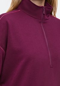 Pullover in pile viola con colletto alto, chiusura a mezza zip e cuciture rosa a contrasto sulle spalle. Tessuto morbido e textured.