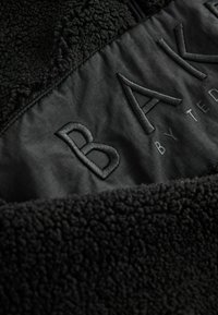 Zwarte fleecejas met een textuurmateriaal en een gladde stoffen paneel met geborduurd logo in een contrasterende kleur.