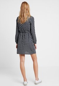 Robe noire avec des motifs en forme de goutte blanche, manches longues, taille élastique et une jupe au-dessus des cuisses. Assortie à des baskets blanches.
