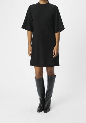 Personne portant une robe noire mi-longue avec des manches arrivant au coude et des bottes noires montantes à bout pointu sur un fond blanc.