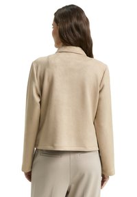 Beżowa kurtka z imitacji zamszu z kołnierzem z klapą i długimi rękawami. Gładka faktura o skróconej długości. Stylizowana z beżowymi spodniami.