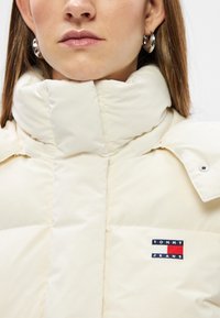 Kremowa kurtka puchowa z wysokim kołnierzem, z teksturowaną powierzchnią i małym, wielokolorowym logo "TOMMY JEANS" na piersi.