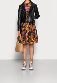 Svart läderjacka i kort modell över en blommig klänning med gula, rosa och gröna mönster. En beige handväska och tan färgade högsneakers fulländar looken.