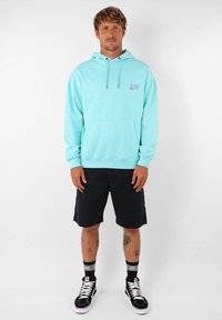 Pull bleu clair avec poche avant, cordons de serrage réglables et détails brodés. Associe avec un short noir et des baskets montantes.