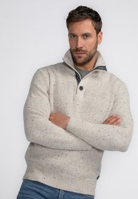 Pull en tricot beige clair avec un motif texturé, présentant un col montant, un plastron à deux boutons et des poignets et un ourlet côtelés.