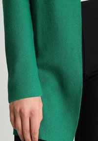 Veste en tricot verte avec des manches longues, une surface texturée et une ouverture à l'avant. Tissu noir visible en dessous. Design simple et sans embellissement.