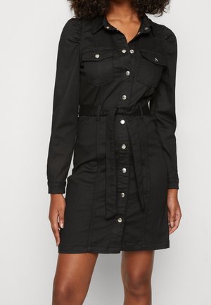Femme portant une robe noire boutonnée sur le devant, à manches longues, avec des poches poitrine et une ceinture nouée à la taille.