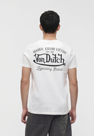 Mann trägt ein weißes Von Dutch T-Shirt mit dem Aufdruck „Original Kustom Kulture 1929-1992 Legendary Brand“ auf dem Rücken, kombiniert mit dunkler Hose.