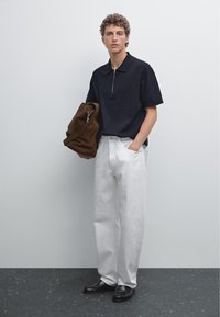 Svart polo med dragkedja, vita vida byxor och svarta skor. Modellen håller en brun mockaväska. Enkel bakgrund, neutrala toner.
