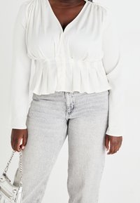 Blusa blanca de manga larga con escote en V y hombros fruncidos, que presenta un dobladillo peplum. Combinada con unos jeans de talle alto en gris claro y un bolso metálico.