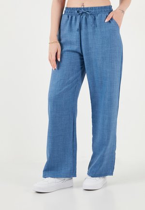 Žena v voľných modrých ľanových nohaviciach s elastickým pásom a bielych teniskách, stojaca s jednou rukou v vrecku na bielom pozadí.