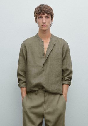 Une jeune personne aux cheveux ébouriffés porte une chemise ample vert olive et un pantalon assorti, se tenant devant un fond doux et clair gris.