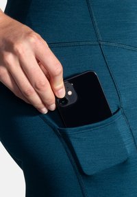 Turkoosiset legginsit, joissa on tiukka istuvuus, sisältävät sivutaskun, johon on sijoitettu musta älypuhelin. Kangas on sileä ja siinä on näkyviä ompelutietoja.