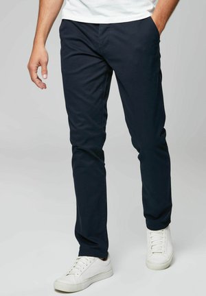 Chinos de algodón azul marino con corte ajustado, bolsillos frontales y pasadores para cinturón, combinados con zapatillas blancas. Textura suave, sin patrones visibles.