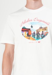 Balta medvilninė marškinėlių suapvalinta grafika vaizduojanti Berlyną, su spalvingais vaizdais, viršuje raudonas užrašas "Adidas Originals" ir apačioje "BERLYNAS".
