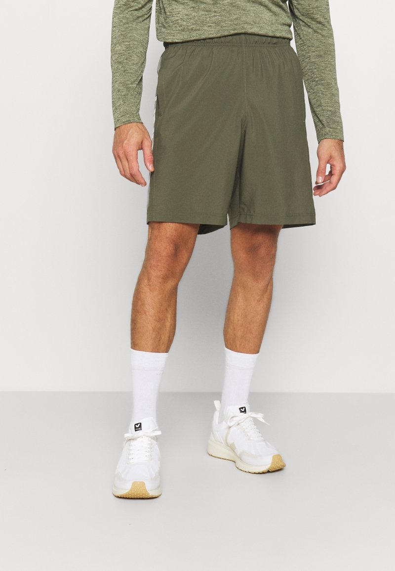 Man som bär olivgröna träningsoveraller, vita ankelsockor och vita sneakers, stående mot en enfärgad vit bakgrund.