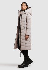 Dolga, svetlo bež puhovka z velikim kapuco, z zadrgami na žepih in črnimi zadrgami. Oblečena čez črne hlače in krepkimi črnimi škornji.