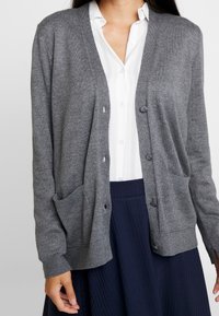 Cardigan gris de tejido de punto suave, con escote en V, cierre de botones, puños acanalados y dos bolsillos frontales. Llevado sobre una camisa blanca.