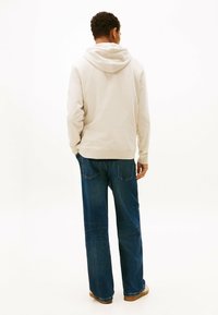 Sudadera beige clara con capucha ajustable, combinada con unos vaqueros de pierna recta azules. Textura de tela suave, con detalles de costura mínimos.