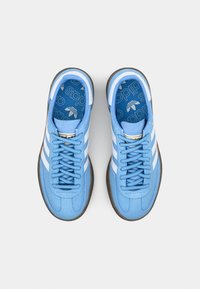 Light blue sneakers featuring white stripes, round laces, and a textured brown rubber sole. The interior has a patterned insole with a logo.