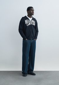 Ung mann står med hendene i lommene, iført svart Billionaire Boys Club hettejakke med glidelås, hvit skjorte, løstsittende blå jeans og svarte joggesko.