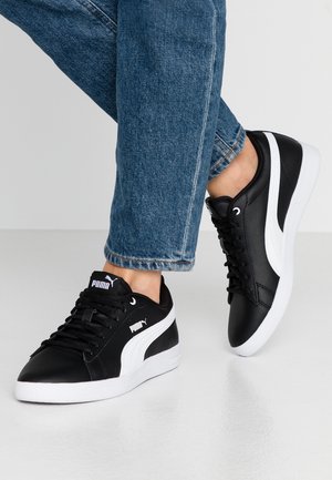 Schwarze Ledersneaker mit weißen Akzenten und Logo, featuring einer abgerundeten Zehe, flacher Sohle und Schnürdesign. Getragen mit blauen Jeans.