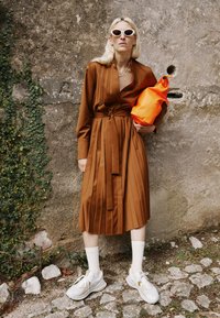 Braunes plissiertes Kleid mit einem gegurteten Taillenbereich, kombiniert mit weißen Turnschuhen und knielangen Socken. Eine leuchtend orangefarbene Tasche sorgt für einen Farbtupfer.