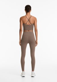 Braun Sport-BH mit gekreuzten Trägern und passender hochgeschnittener Leggings. Glatter, dehnbarer Stoff mit minimalem Branding. Schlichte weiße Sneakers.