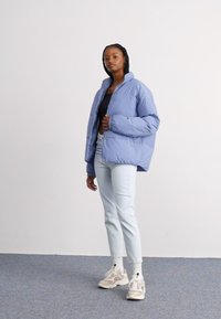 Light blue puffer jacket with a high collar, featuring a smooth texture. Paired with light denim jeans and beige sneakers.