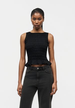 Γυναίκα με πλεξούδες, φορώντας μαύρο τοπ χωρίς μανίκια με σούρες και βολάν στο τελείωμα, καθώς και σκούρο τζιν ψηλόμεσο, στέκεται μπροστά σε απλό φόντο.