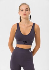 Top deportivo morado corto con tirantes gruesos, escote redondeado, tejido texturizado y un logo negro en el centro. Combinado con leggings de talle alto a juego.