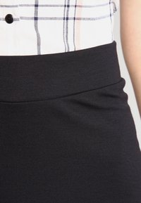 Jupe en tissu extensible noir avec une large taille, texture lisse et design minimal. Associée à une chemise à carreaux en haut.