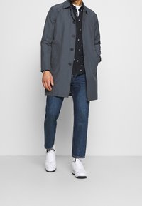 Cappotto grigio leggero con chiusura a bottoni, indossato sopra una camicia nera a fantasia, abbinato a jeans blu e sneaker bianche.