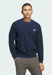 Mørkeblå hettegenser med lange ermer, rund hals og ribbede mansjetter. Har en hvit Adidas-logo på brystet og strukturert stoff.