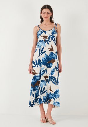 Robe maxi florale présentant des motifs de feuilles bleues et brunes sur un fond crème, avec des fines bretelles et une coupe décontractée, fabriquée en tissu léger.