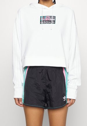 Personne portant un sweat à capuche court blanc Adidas avec logo et un short Adidas noir avec des rayures turquoise et roses sur les côtés.