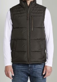 Olivgrüne Steppweste mit hohem Kragen, Front-Reißverschluss, Seitentaschen und horizontalen Nähten. Wird über einem weißen Langarmshirt getragen.