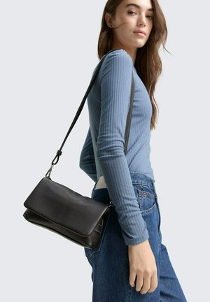 Borsa a tracolla in pelle nera testurizzata con patta pieghevole, indossata sulla spalla, abbinata a una maglietta a maniche lunghe blu a costine e jeans.