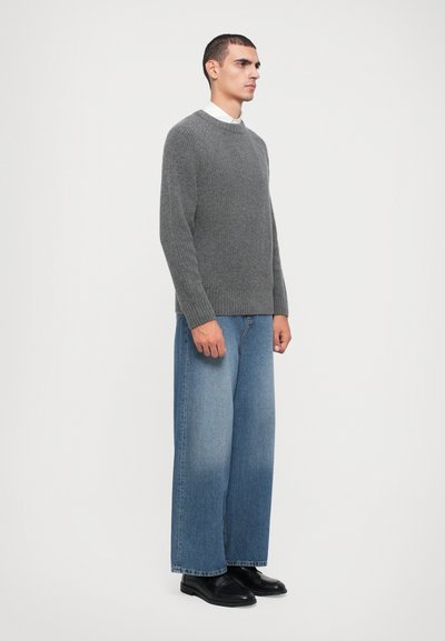 Pull en maille gris avec un ourlet côtelé, associé à un jean en denim bleu clair à jambe large. Le modèle porte des chaussures noires. Design simple et épuré ; sans motifs.