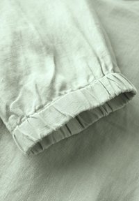 Tissu en lin vert clair avec un détail à manche froncée. La texture apparaît douce et légèrement froissée, accentuant un look décontracté.