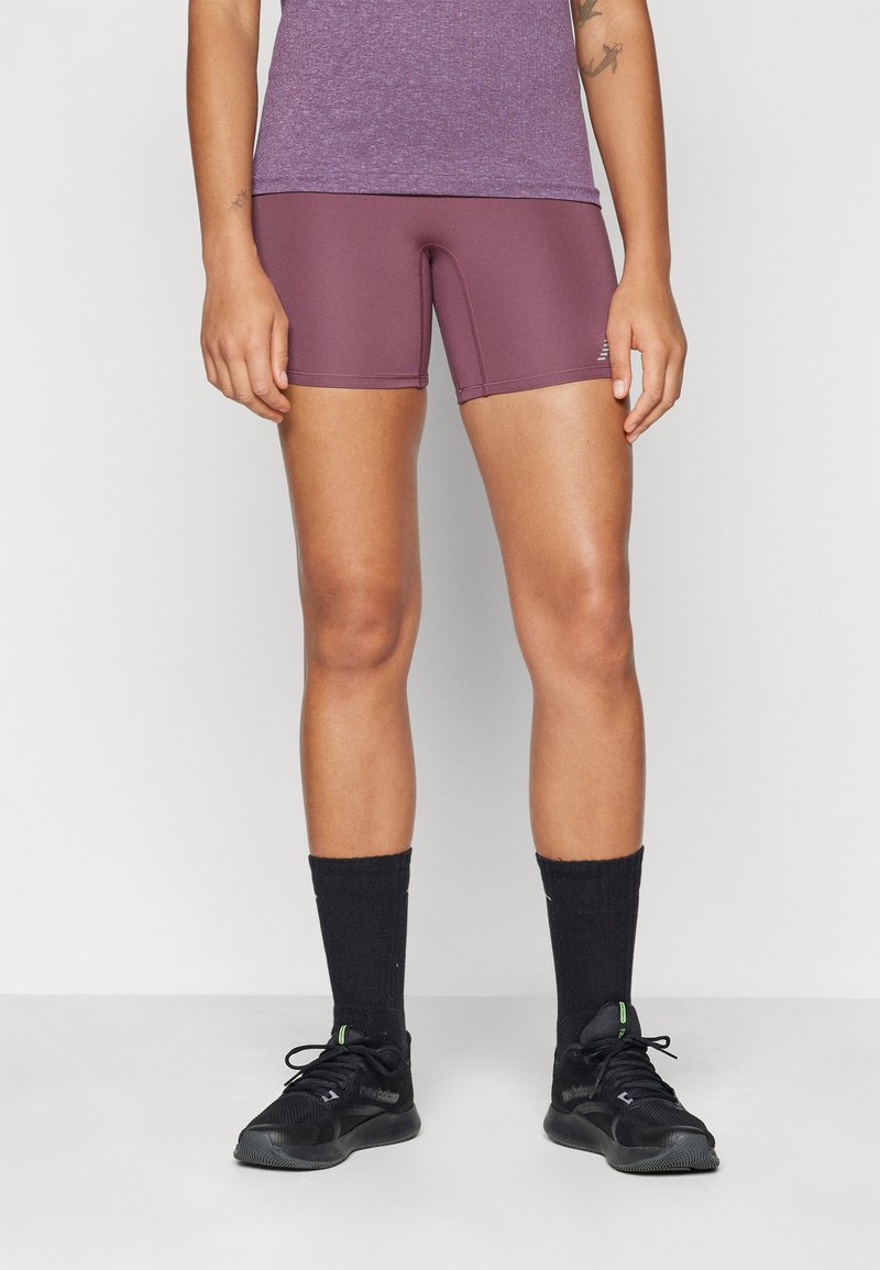 Shorts de sport violets en tissu extensible, avec une coupe ajustée. Portés avec des chaussettes de cheville noires et des chaussures de sport noires.